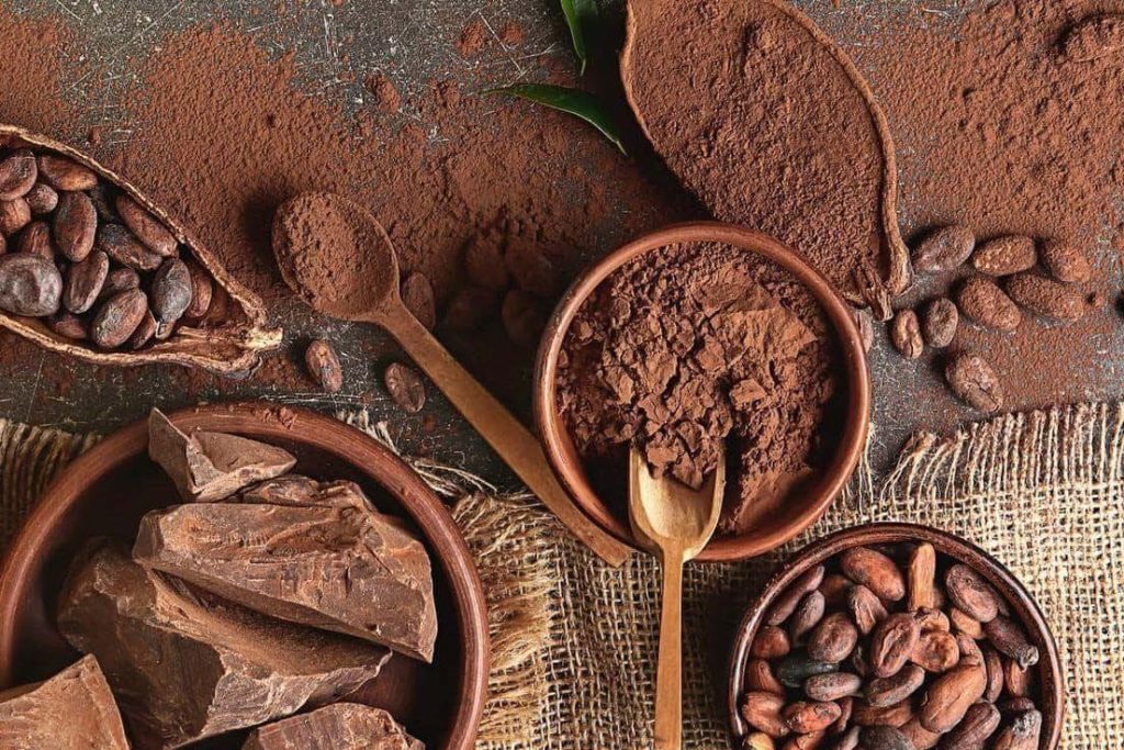 Bàn gỗ với bột cacao, hạt cacao, và thanh sô cô la thủ công – minh họa sự khác biệt giữa sô cô la nguyên chất và công nghiệp.