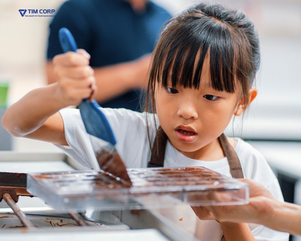Bé gái tham gia trải nghiệm F&B, đổ socola vào khuôn trong một lớp học nấu ăn cho trẻ em.