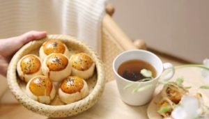 Giỏ bánh nướng vàng óng rắc mè đen bên tách trà nóng, đặt trên bàn gỗ tạo cảm giác ấm cúng và thư giãn.