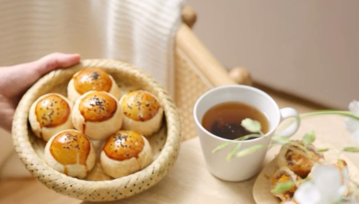 Giỏ bánh nướng vàng óng rắc mè đen bên tách trà nóng, đặt trên bàn gỗ tạo cảm giác ấm cúng và thư giãn.